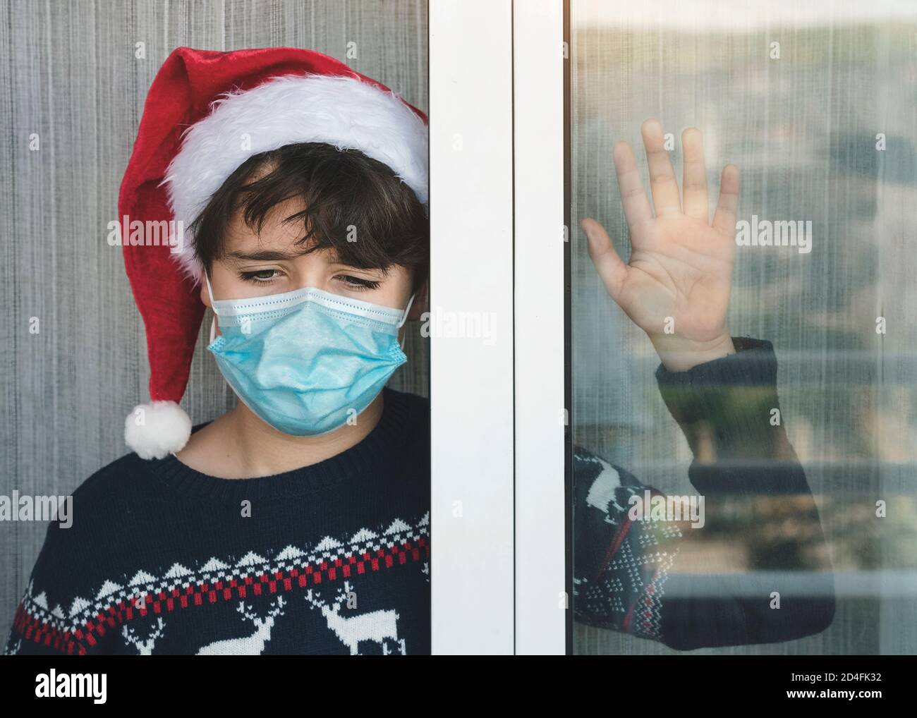 Triste bambino con maschera medica che indossa cappello Babbo Natale aspettare Per babbo natale accanto a una finestra di Natale da La pandemia di Coronavirus Foto Stock