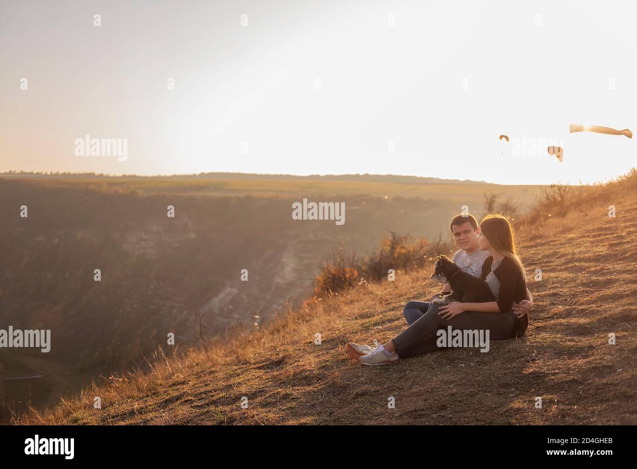 Seduto su un canyon, guarda i parapendio che volano nel cielo al tramonto. Viaggio con animali domestici. Giovane famiglia con un cane schnauzer viaggio. Moldavia, Vecchia Orhey Foto Stock