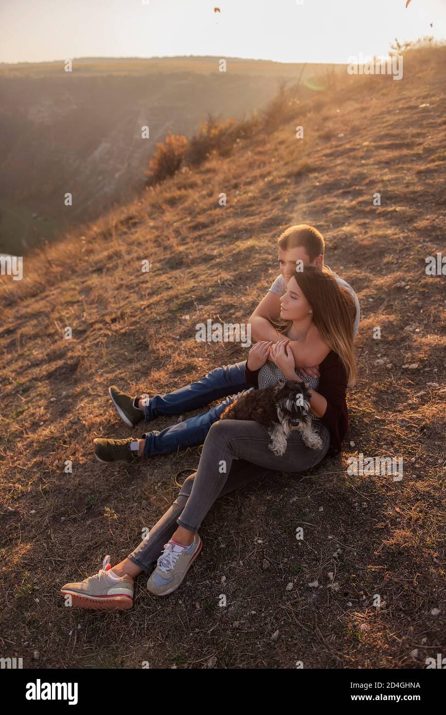 Un giovane abbracca una bella ragazza da dietro, la coppia ammira il tramonto dalla cima della collina. Felice famiglia viaggia con un cane schnauzer. Foto Stock