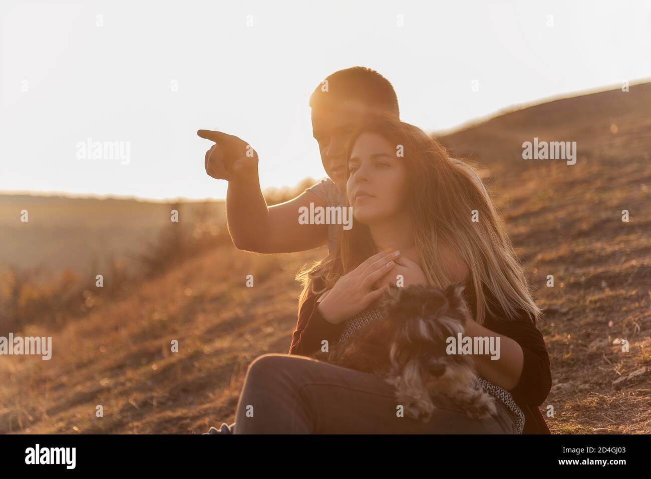 Un giovane abbracca una bella ragazza da dietro, la coppia ammira il tramonto dalla cima della collina. Felice famiglia viaggia con un cane schnauzer. Foto Stock