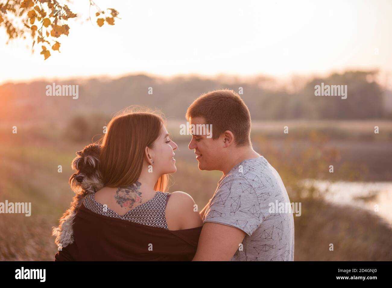 Una coppia in abbracci d'amore, bacia nei raggi del sole d'autunno, con le spalle girate verso la macchina fotografica, tenendo una Schnauzer tra le braccia. Bella ragazza Foto Stock