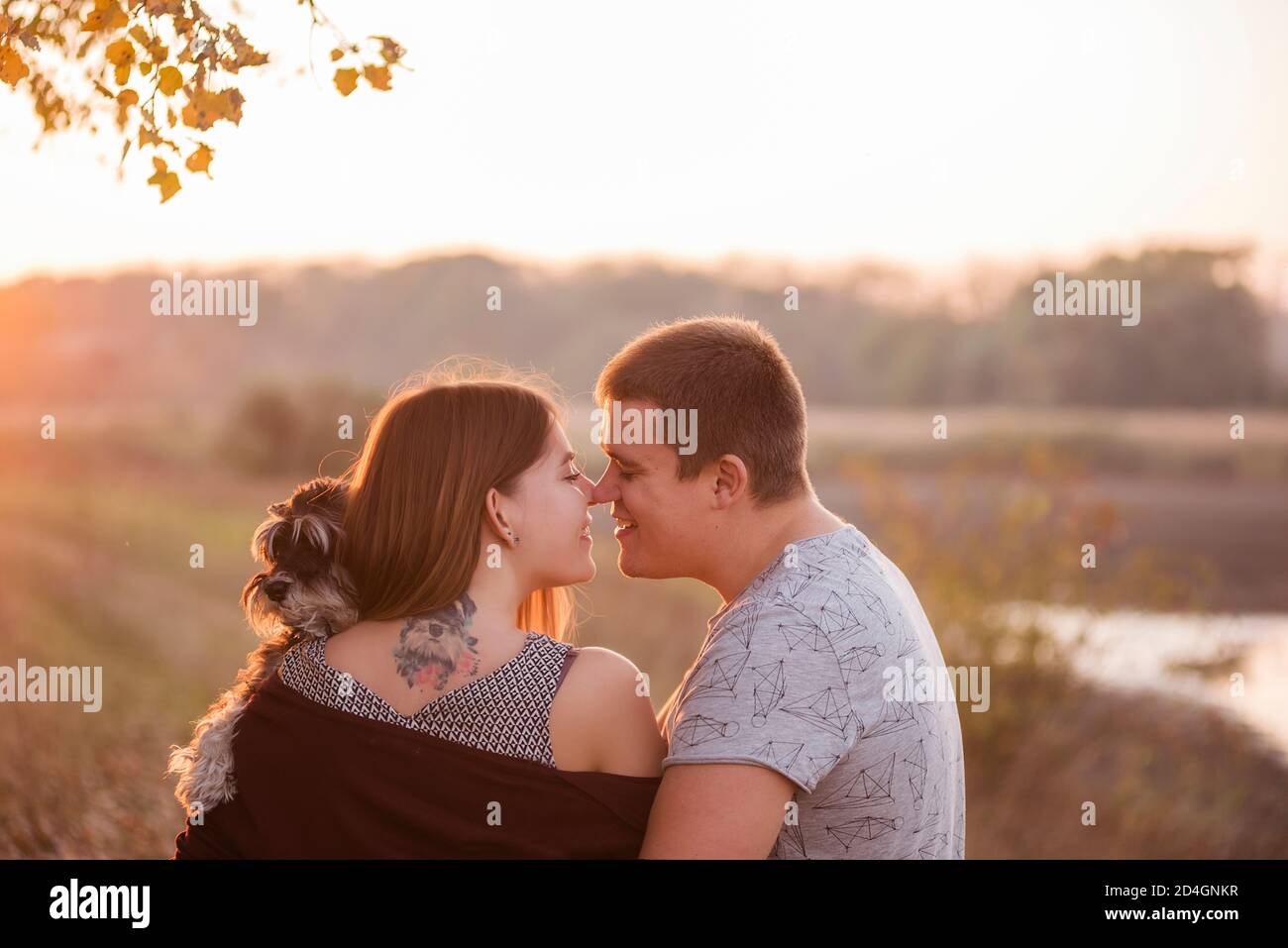 Una coppia in abbracci d'amore, bacia nei raggi del sole d'autunno, con le spalle girate verso la macchina fotografica, tenendo una Schnauzer tra le braccia. Bella ragazza Foto Stock