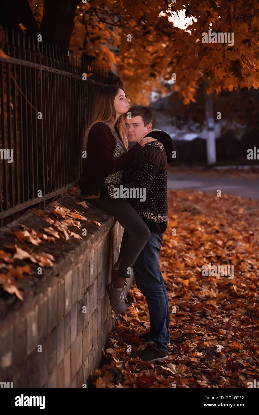 Una giovane coppia di adolescenti si siede vicino alla recinzione sotto il fogliame arancione di un albero nella sera d'autunno. Gli amanti abbraccio, guardarsi, bacio. Foto Stock