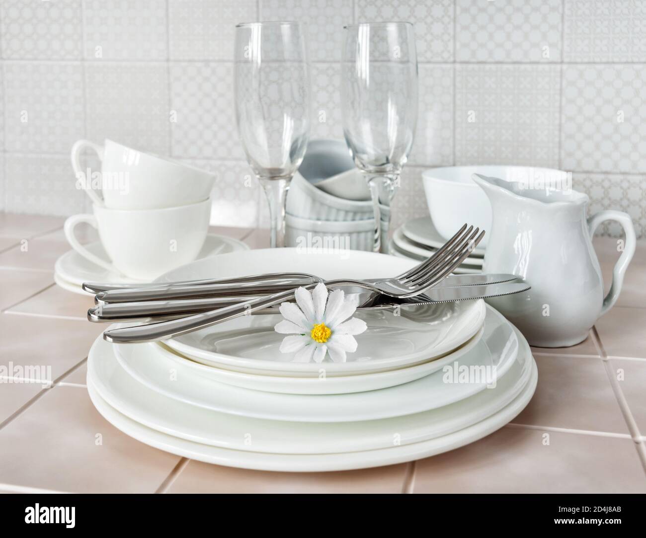 Pila di piatti in porcellana bianca, tazzine da tè e bicchieri da vino sul tavolo da cucina sullo sfondo di un grembiule in ceramica Foto Stock