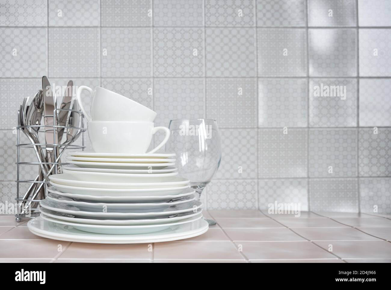 Pila di piatti in porcellana bianca, tazzine da tè, un bicchiere di vino, nonché cucchiai e forchette nel supporto per piatti sul tavolo da cucina contro il backgrou Foto Stock