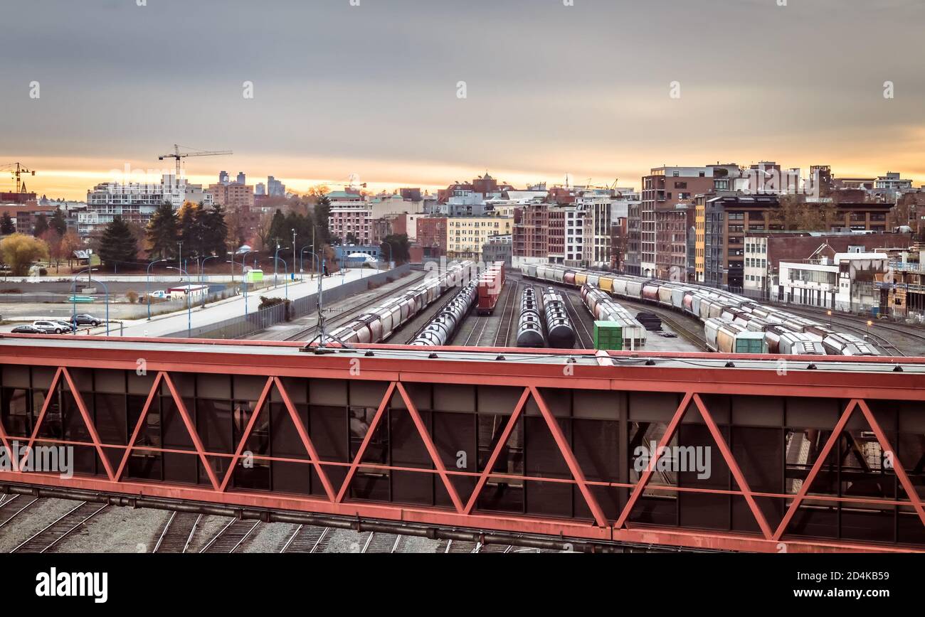 Treni merci in città. Alba scena con paesaggio urbano residenziale grandi gru di costruzione. La passerella pedonale per il mare è in primo piano. Va Foto Stock