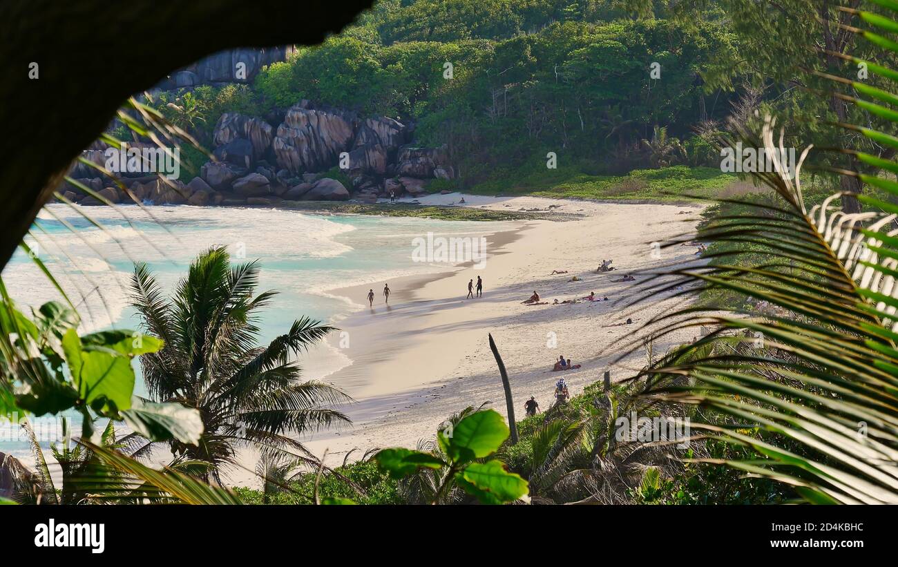 Vista attraverso la vegetazione con turisti prendere il sole sulla splendida spiaggia tropicale Grand Anse nel sud dell'isola di la Digue, Seychelles. Focus sulla spiaggia. Foto Stock