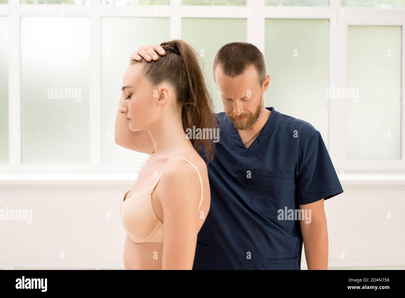 Uomo bearded in uniforme che tocca la testa della donna in intimo durante la sessione di osteopatia in clinica contemporanea Foto Stock