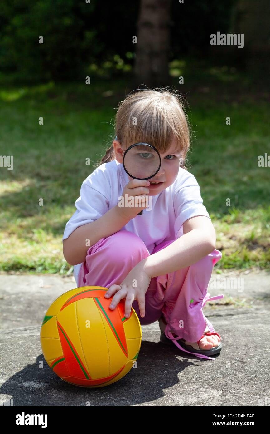 Piccola ragazza felice intelligente, bambino che guarda attraverso una lente d'ingrandimento, capretto con una lente e un gioco di pallacanestro nel giardino esterno. Curioso piccolo gir Foto Stock