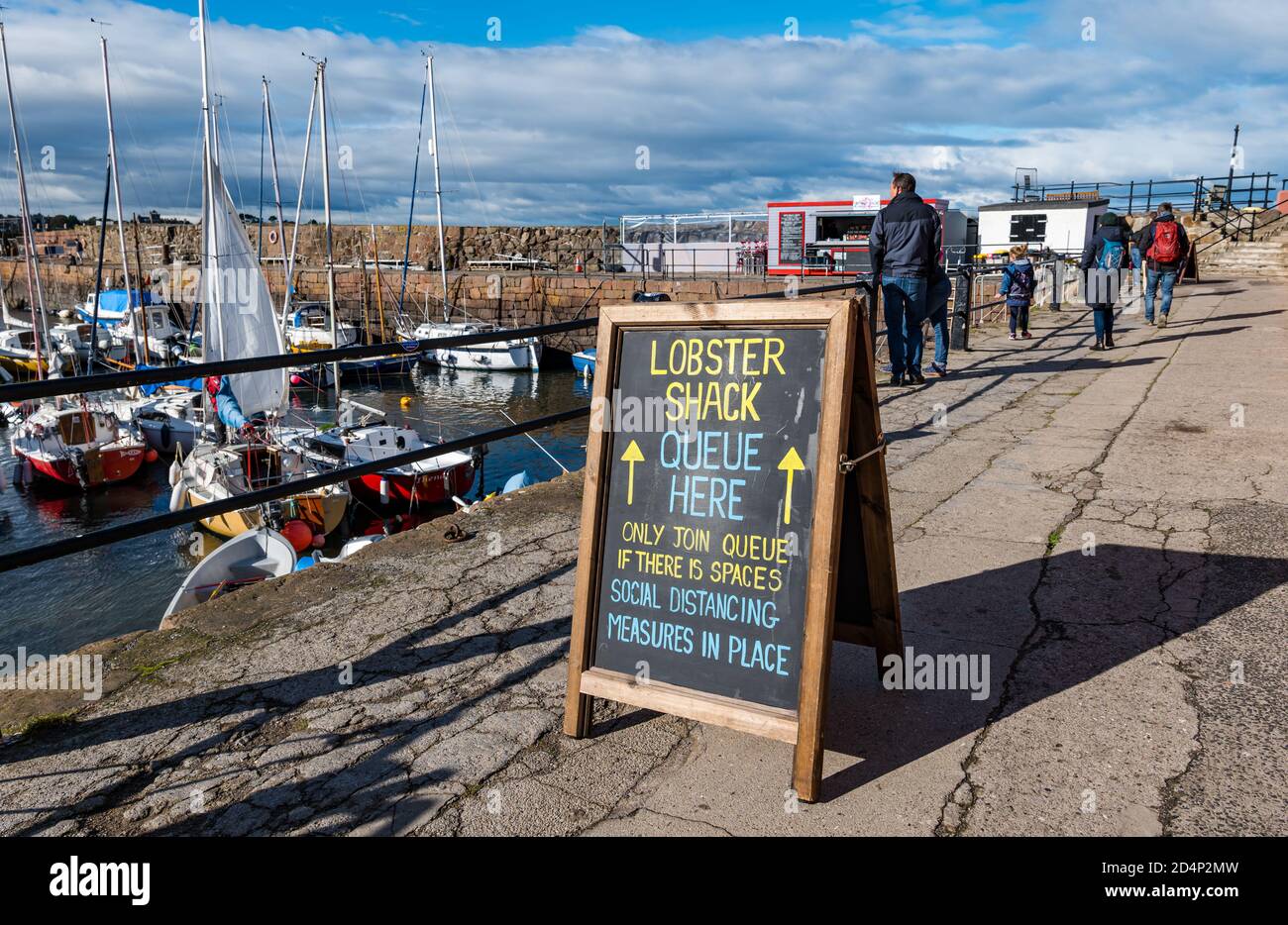 North Berwick, East Lothian, Scozia, Regno Unito, 10 ottobre 2020. UK Weather: Il popolare Lobster Shack attrae i clienti tra le colorate barche a vela nel porto in una giornata ventosa con una bacheca che consiglia ai clienti di mantenere la distanza sociale in coda Foto Stock