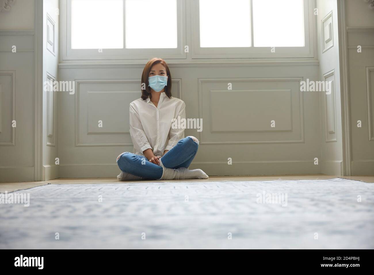 giovane donna asiatica in quarantena a casa con maschera facciale seduta sulle gambe del pavimento incrociate Foto Stock