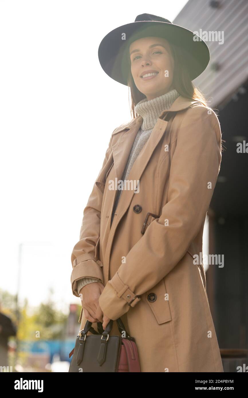 Bella donna con cappello nero, trench beige e pullover grigio che ti guarda Foto Stock
