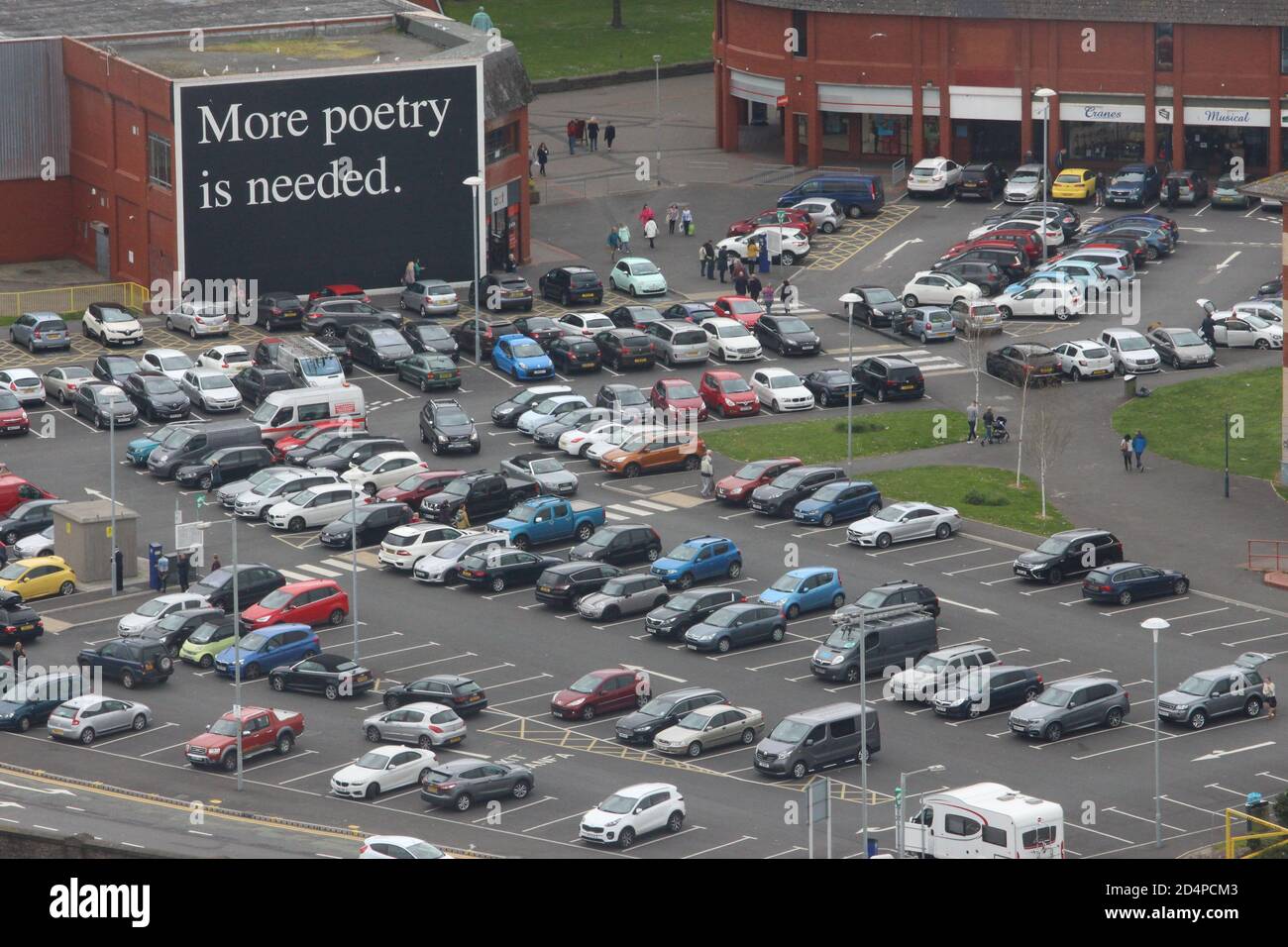 Una fotografia di un parcheggio pieno di veicoli, con una grande parete dipinta con la citazione "più poesia è necessaria". I graffiti dell'arte della Comunità ispirano le parole Foto Stock