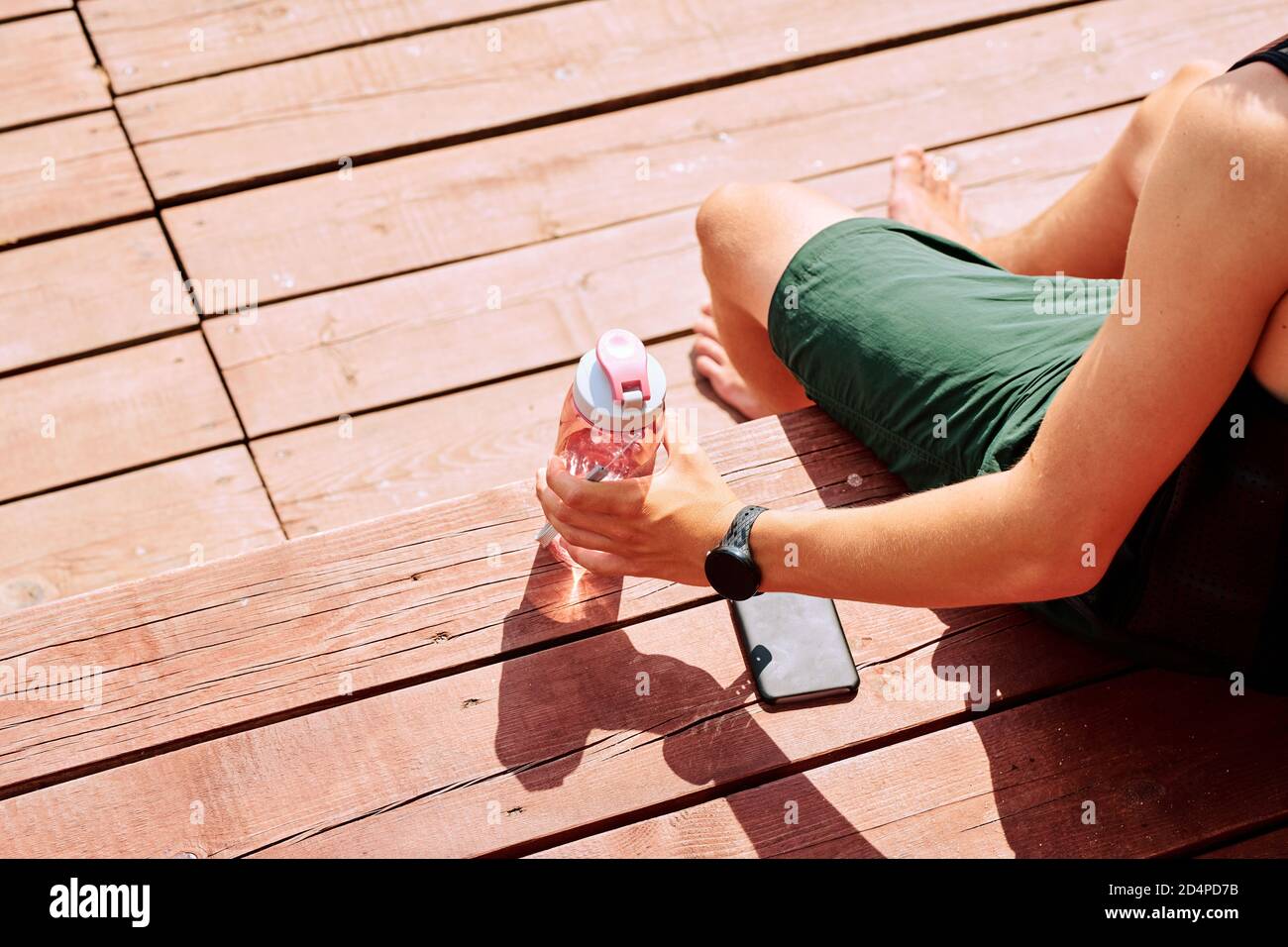 Vista di giovane sportivo in pantaloncini verdi che prendono una bottiglia di acqua da panca di legno Foto Stock
