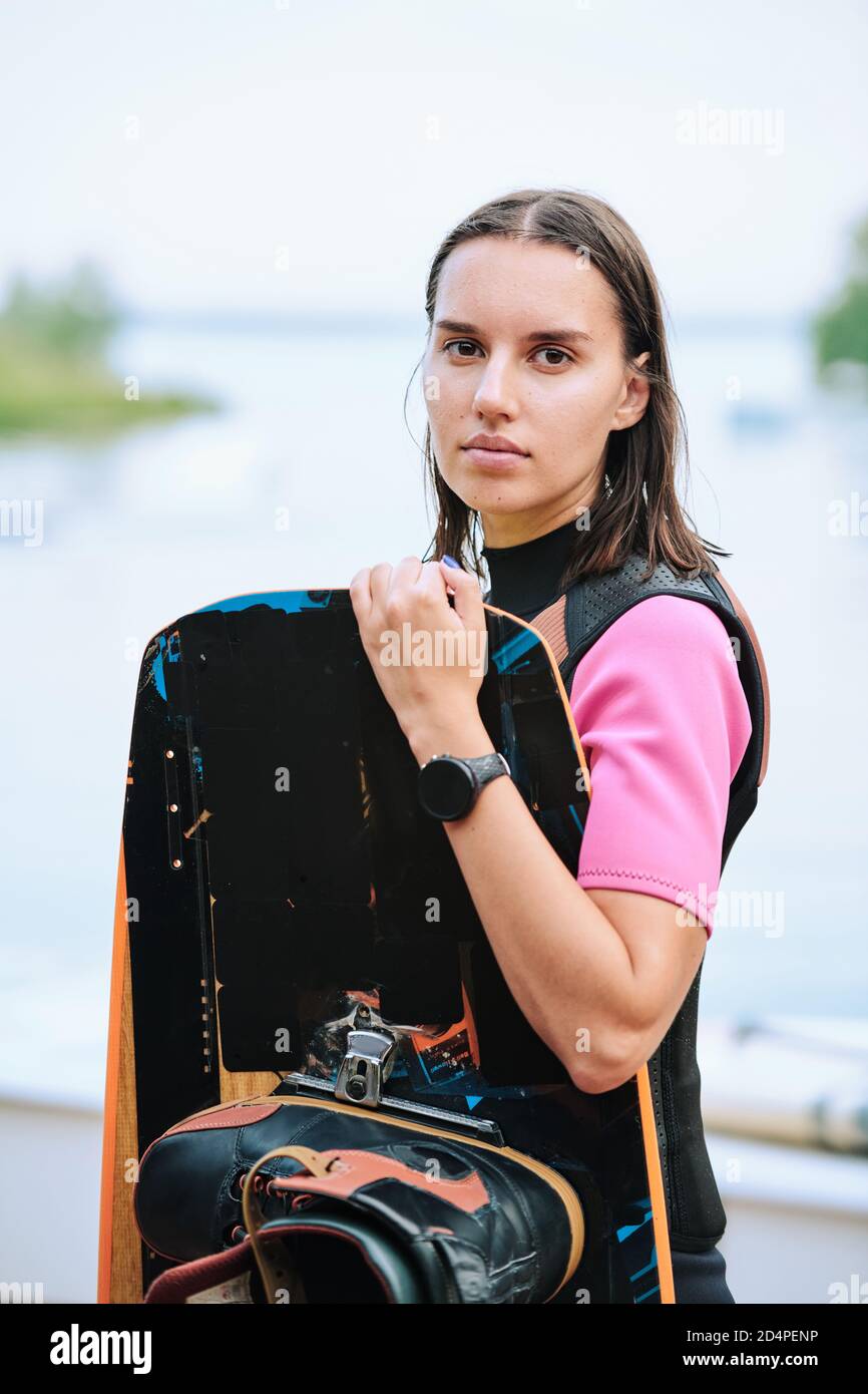 Surfista giovane attivo con capelli scuri che tiene la tavola da surf contro lato acqua Foto Stock