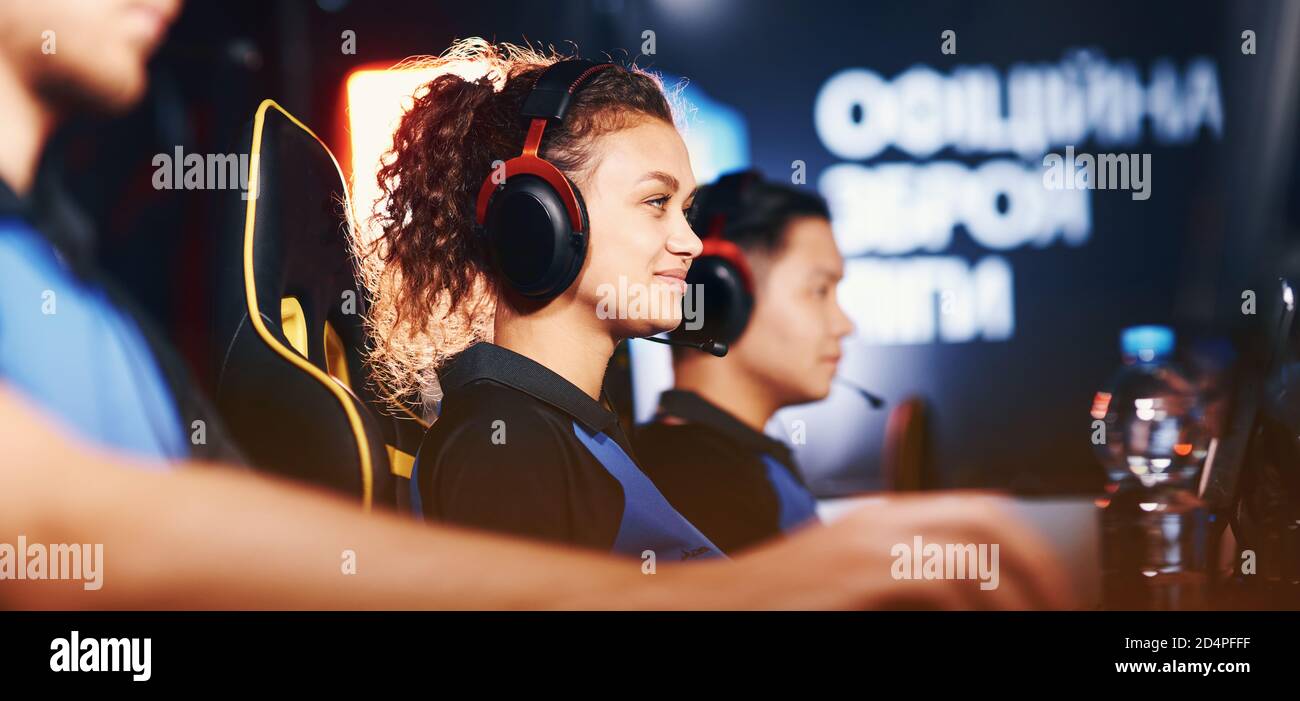 Evento professionale eSport. Vista laterale di una ragazza di razza mista felice, giocatore di cybersport femminile indossando le cuffie che giocano i video giochi online e sorridendo, partecipando con la squadra professionale in torneo Foto Stock