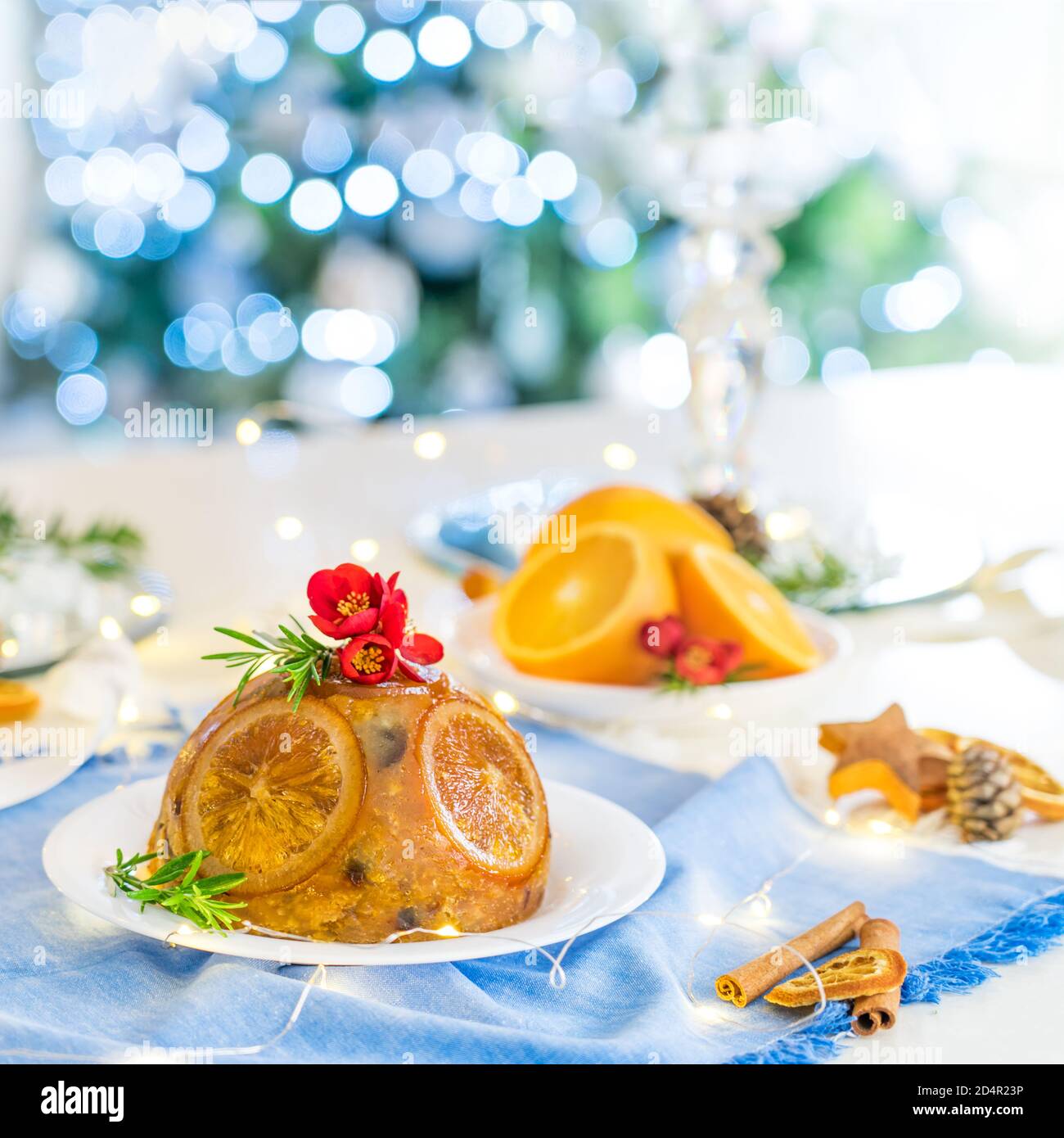 Tradizionale budino di Natale inglese decorato con arance e fiori in un ambiente festoso tavolo con frutta, spezie, luci di albero di Natale su b Foto Stock