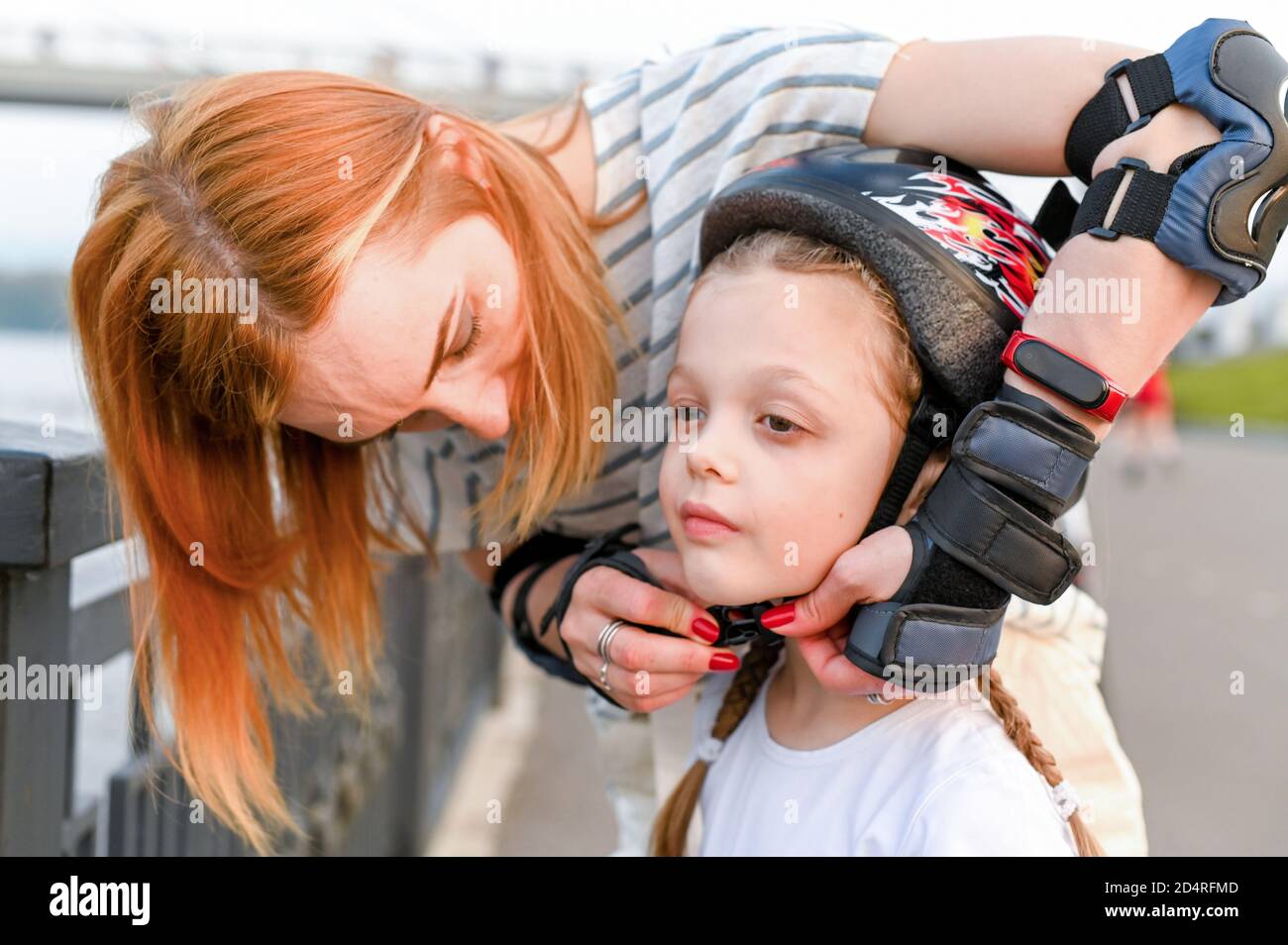 Giovane madre che aiuta la sua piccola figlia a mettere un casco da bicicletta. Bambini che indossano hemet di sicurezza durante il pattinaggio a rotelle Foto Stock
