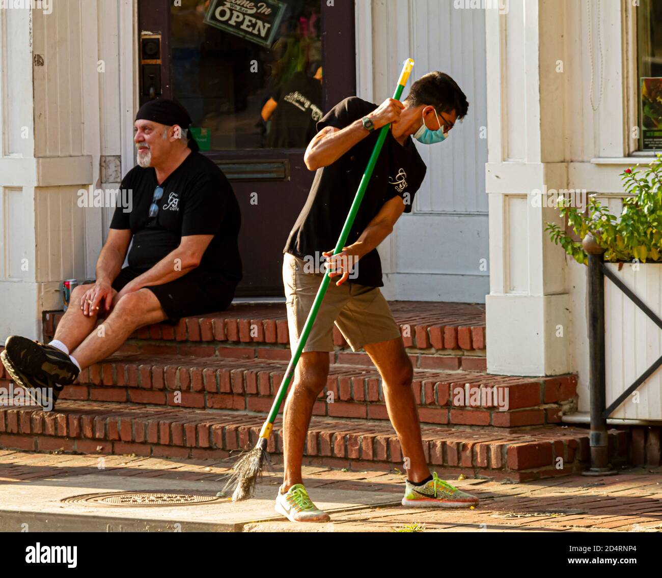Ellicott City, MD, USA 10/07/2020: Un giovane operatore janitorial ispanico che indossa una maschera facciale a causa di COVID sta spazzando il pavimento di fronte al negozio. Il suo c Foto Stock