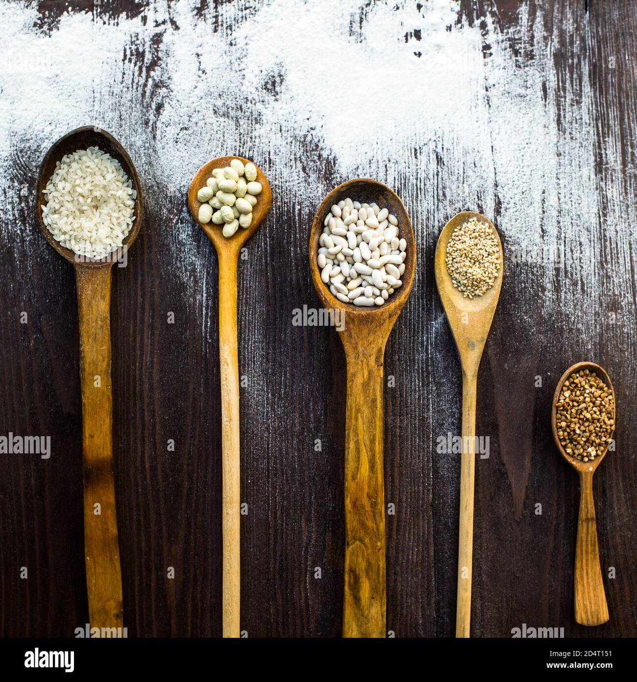 Vista dall'alto di cucchiai di legno sui quali si trovano diversi cereali su un tavolo cosparso di farina. Foto Stock