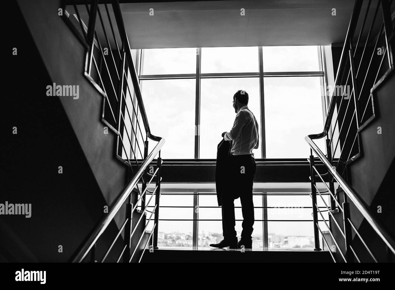 Immagine in bianco e nero dello sposo che guarda fuori dalla finestra e tiene una giacca sulle scale. Foto Stock