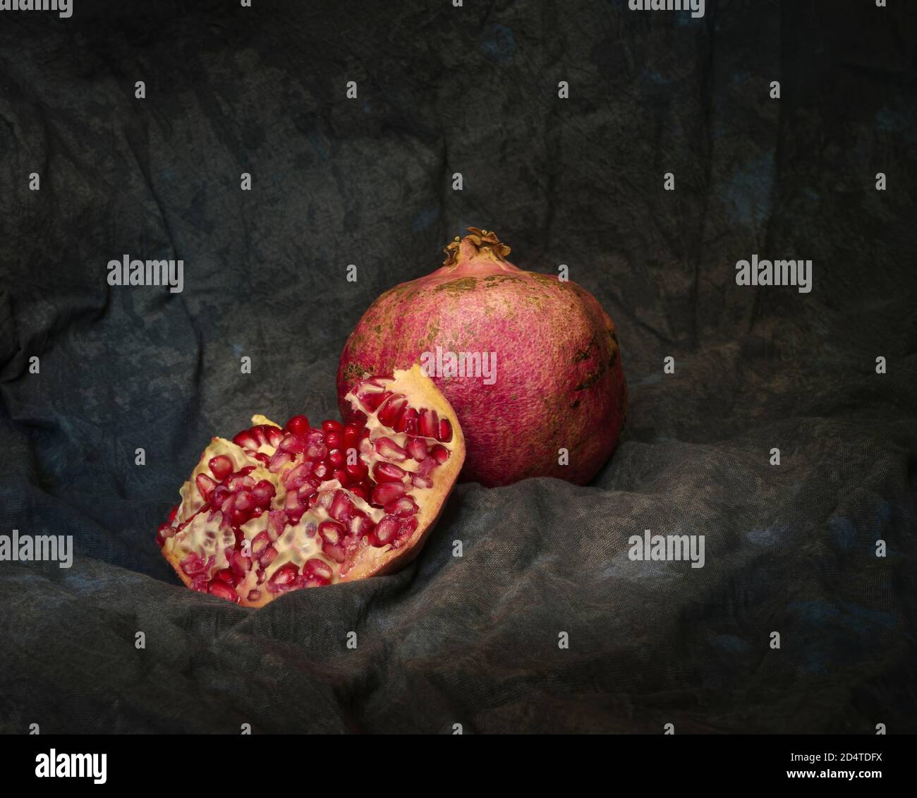 Ancora vita con due melograni coltivati organicamente, uno di loro si divide per vedere l'interno su uno sfondo di tessuto scuro. Foto Stock