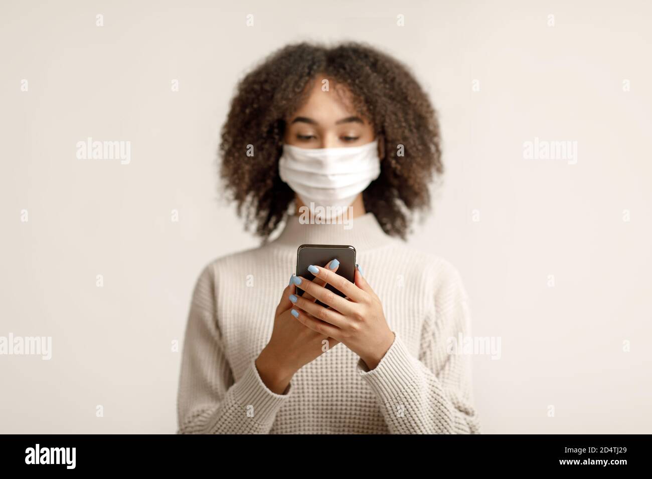 Attraente donna afroamericana in maschera protettiva digitando sullo smartphone e assistenza sanitaria durante la quarantena Foto Stock