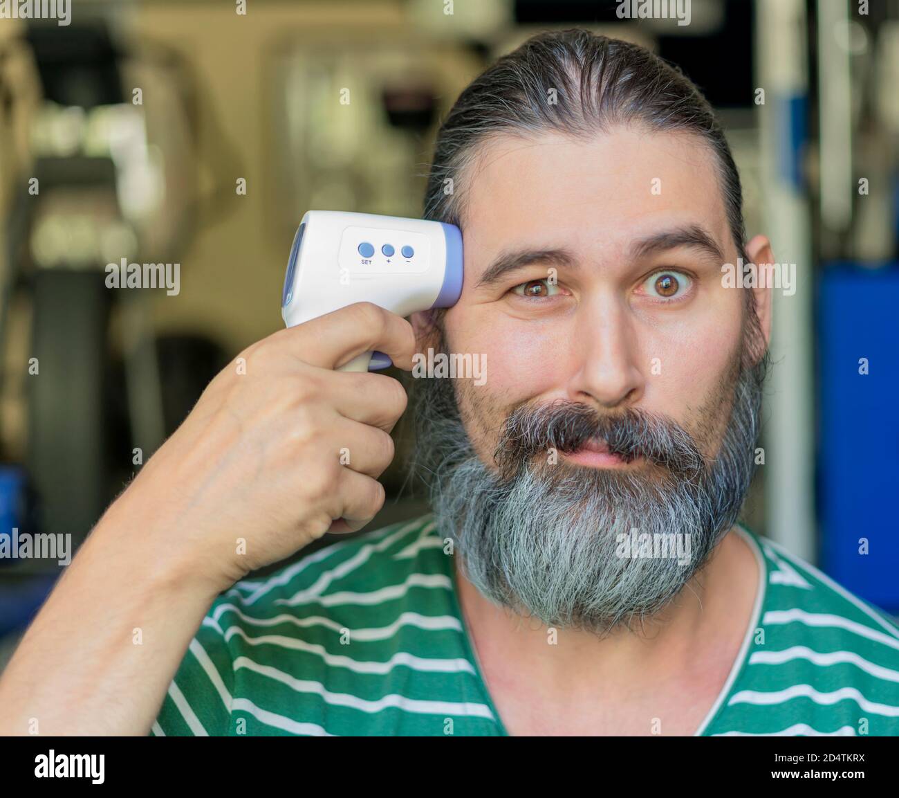 Uomo bearded con i capelli lunghi scuri punta un termometro ad infrarossi al suo viso come se fosse una pistola. Self care, concetto medico e divertente. Foto Stock
