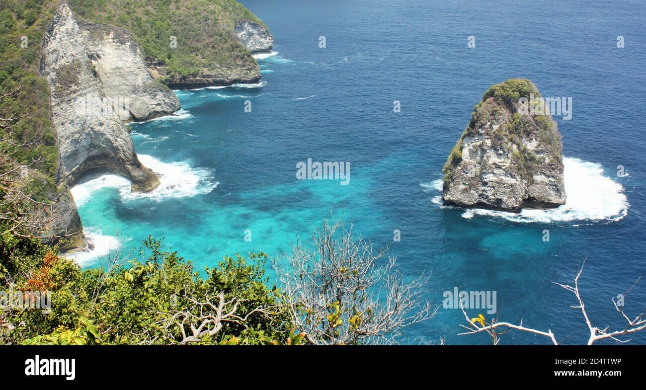 Vista panoramica del mare azzurro turchese e della costa rocciosa con fogliame verde in primo piano sull'isola di Nusa Penida, Bali, Indonesia Foto Stock