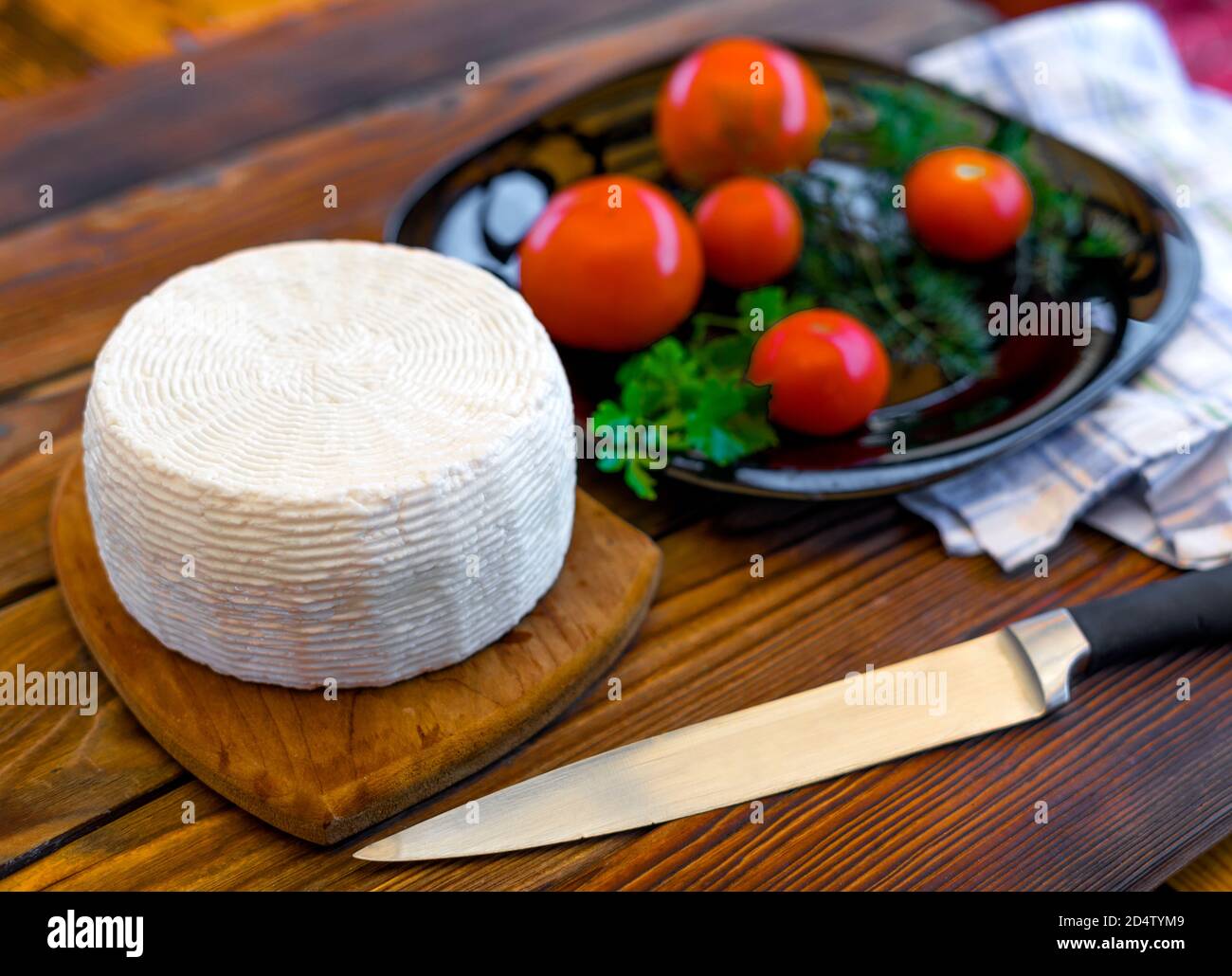 Testa di formaggio su tavola di legno su tavola di legno servita con pomodori e verdure fresche, coltello e asciugamano. Formaggio fatto in casa. Concetto di cibo. Messa a fuoco selettiva. Foto Stock