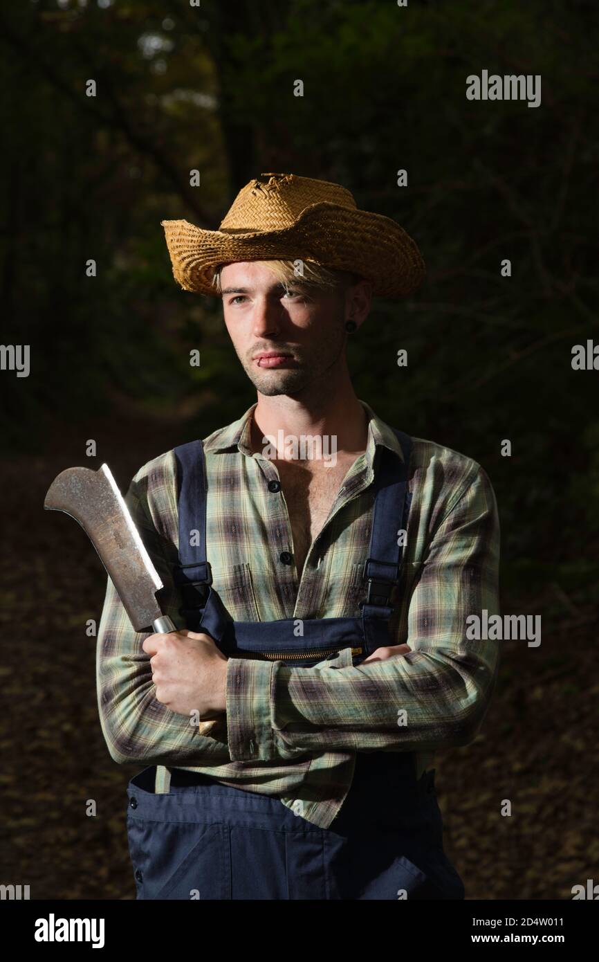 Uomo dai capelli biondi con un falcetto, con dungarees e un cappello di paglia. Foto Stock