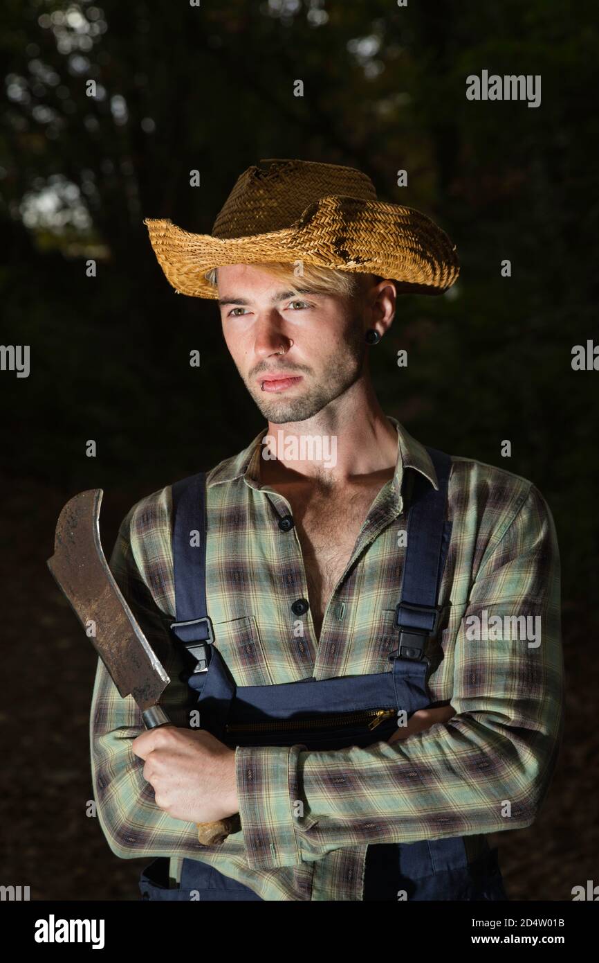 Uomo dai capelli biondi con un falcetto, con dungarees e un cappello di paglia. Foto Stock