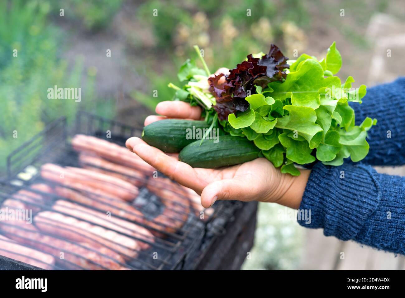 Le mani delle donne tengono insalata ingredienti: Cetrioli, lattuga, basilico. Picnic. Le salsicce vengono cucinate sulla griglia. Messa a fuoco selettiva. Vista ravvicinata. Foto Stock
