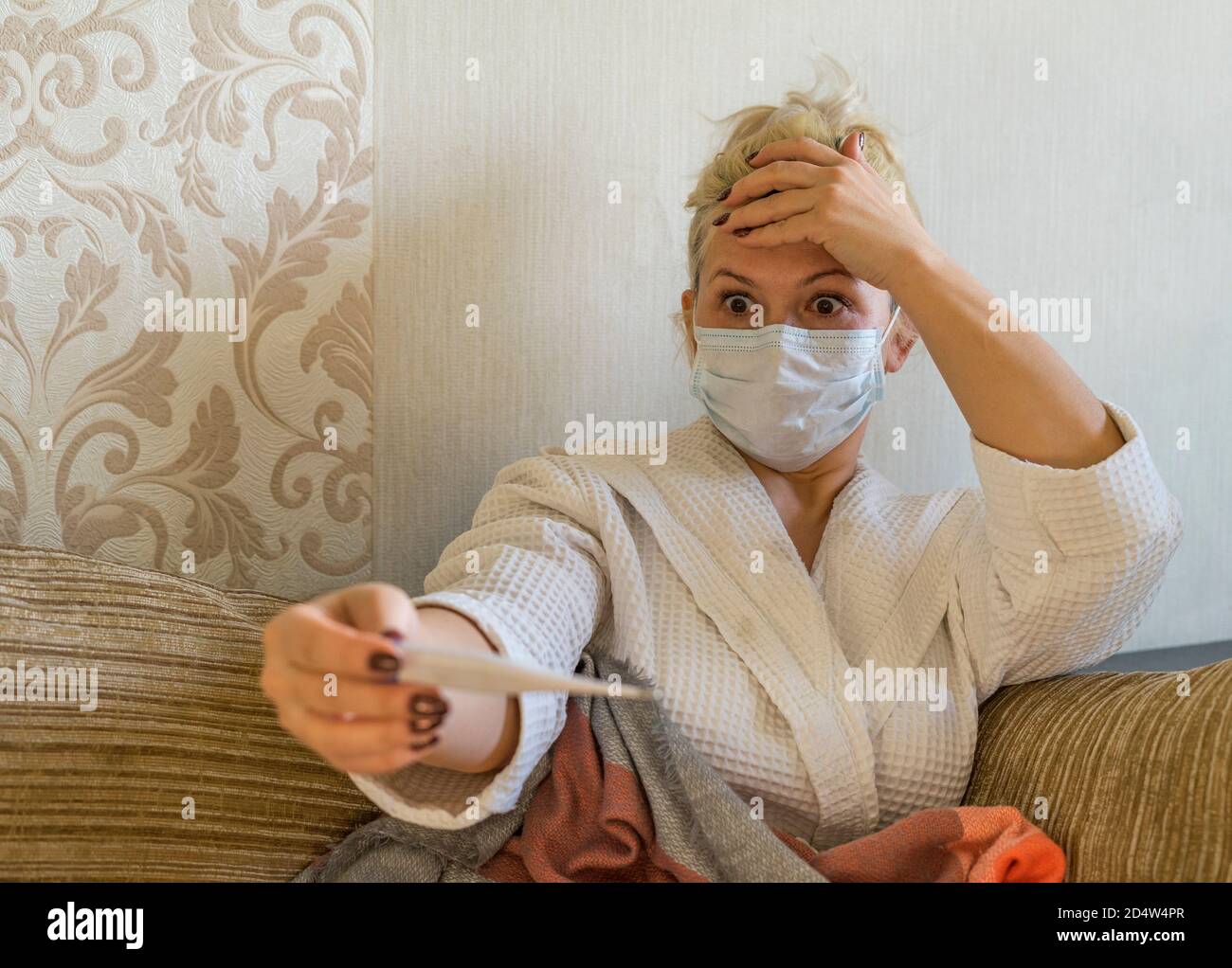 Una donna in una maschera medica si stares largo - eyed ad un termometro. Rimani a casa. Rimani al sicuro. Foto Stock