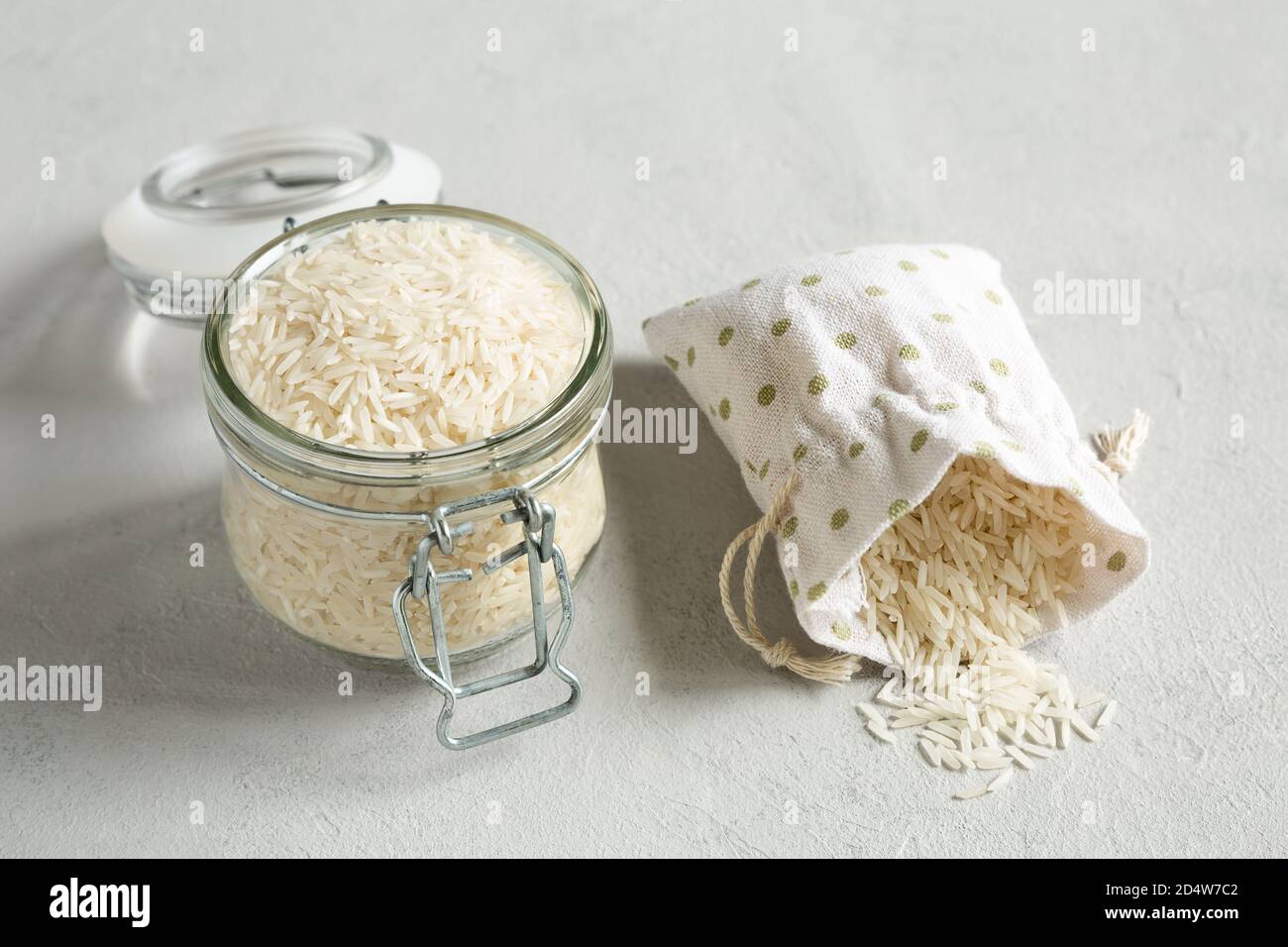 Grano di riso crudo in un vaso di vetro e cereali in un sacchetto di tela su uno sfondo grigio di cemento. Metodi di conservazione di grani. Sano, organico, dietetico Foto Stock