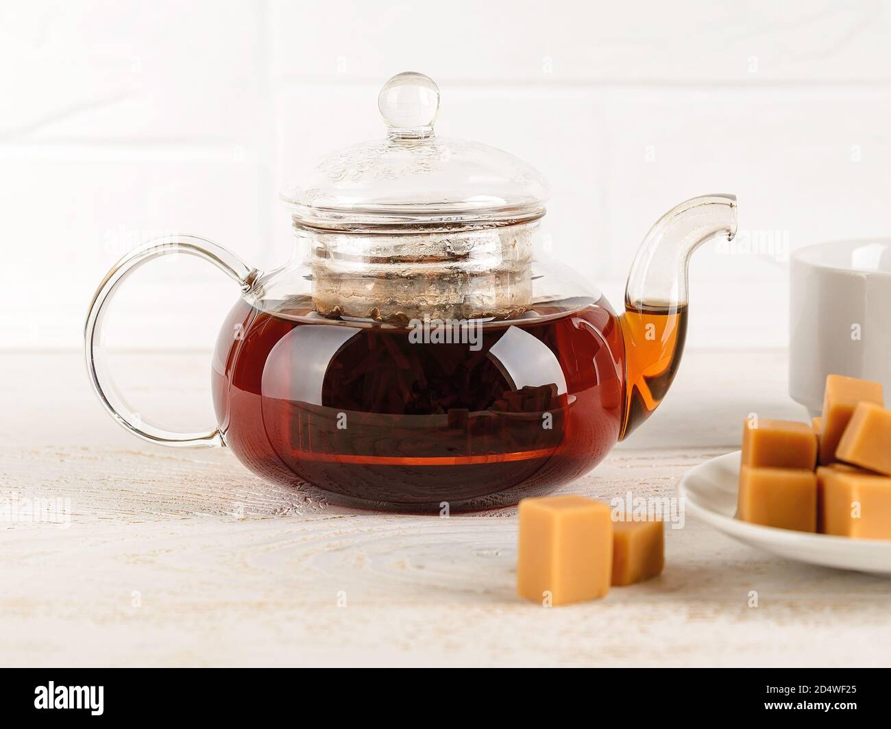 Teiera in vetro trasparente piena di tè nero caldo appena preparato vicino alla tazza e caramello alla vaniglia su un tavolo di legno bianco. Tè bevendo con dolci. Vista frontale Foto Stock