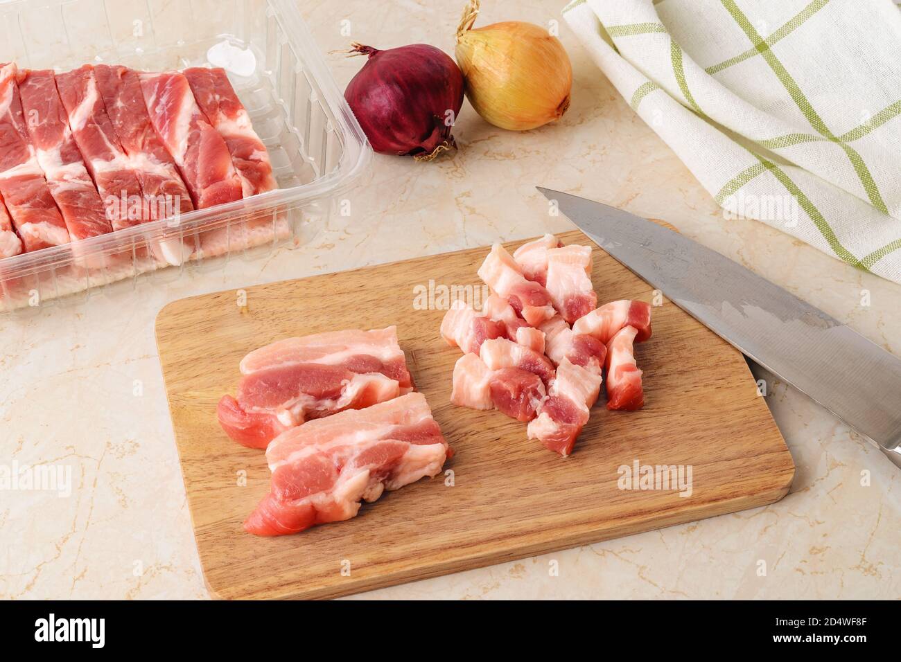 Taglio fresco pancia di maiale crudo in piccoli pezzi su un tagliere di legno con un coltello affilato su un tavolo da cucina. Carne in un contenitore di plastica monouso Foto Stock