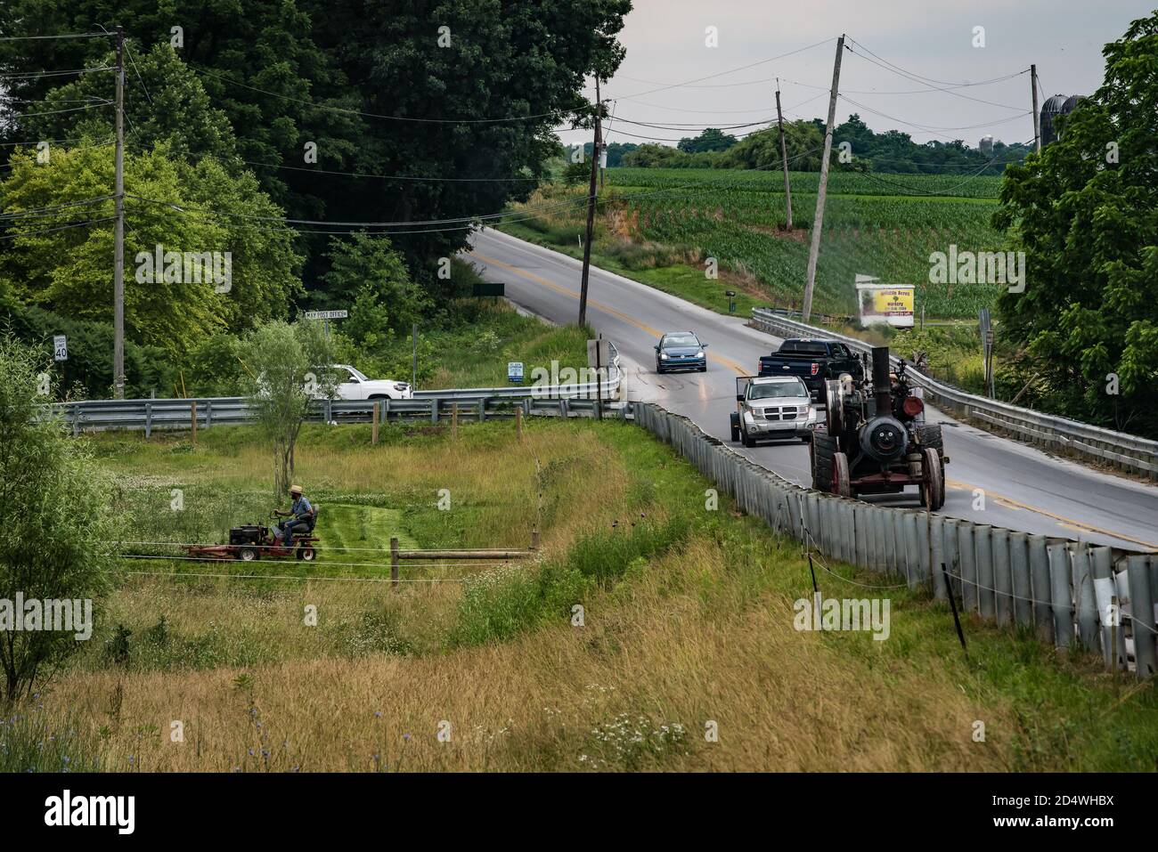 Amish, contea di Lancaster, Pennsylvania. USA antico trattore a vapore rallenta il traffico. L'uomo taglia il giardino con un trattore moderno. Foto Stock