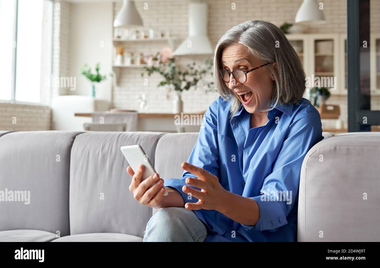 Stupita donna anziana matura che legge il messaggio sullo smartphone a casa. Foto Stock