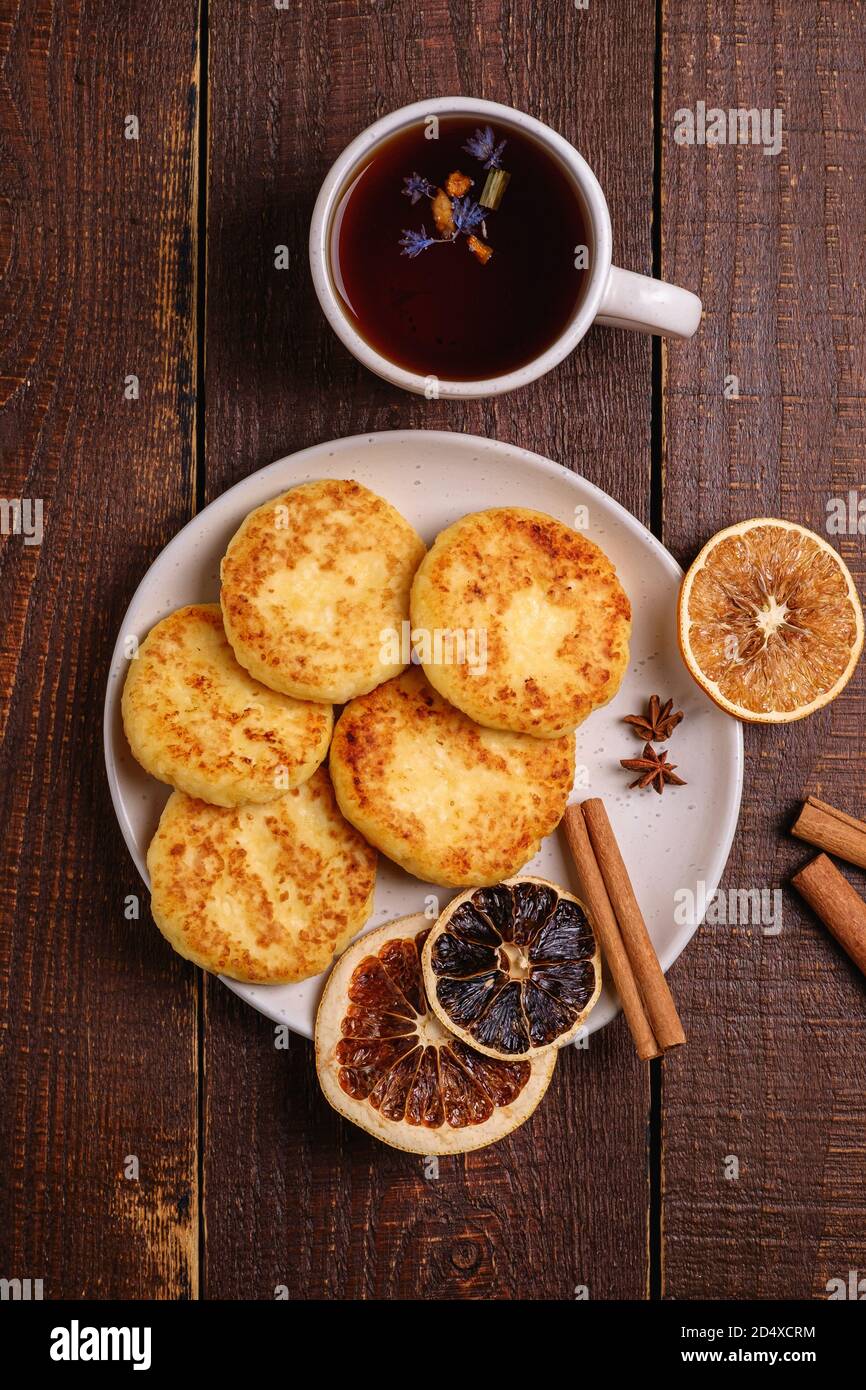 Frittelle di formaggio cottage con tè nero aromatico caldo, umore di prima colazione di Natale con anice, cannella e agrumi secchi su sfondo di legno, vista dall'alto Foto Stock