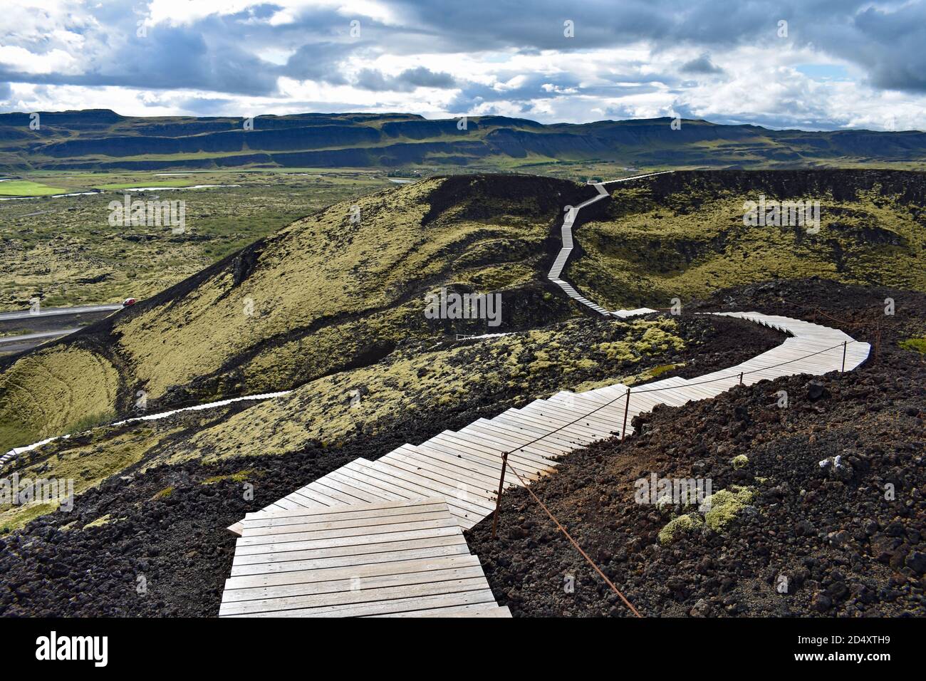 Una passerella in legno si snoda intorno al bordo del Cratere del Vulcano Grabrok nella Valle di Nordurardalur, nell'Islanda occidentale. Il muschio verde copre la roccia nera. Foto Stock