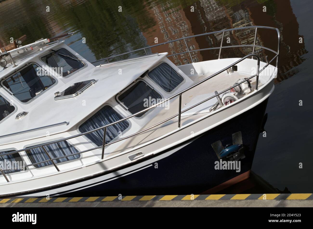 Ormeggiata a riva è una barca da diporto con ponte bianco con ringhiera in metallo, tende blu disegnate dietro le finestre delle cabine e scafo nero su una superficie d'acqua scura Foto Stock