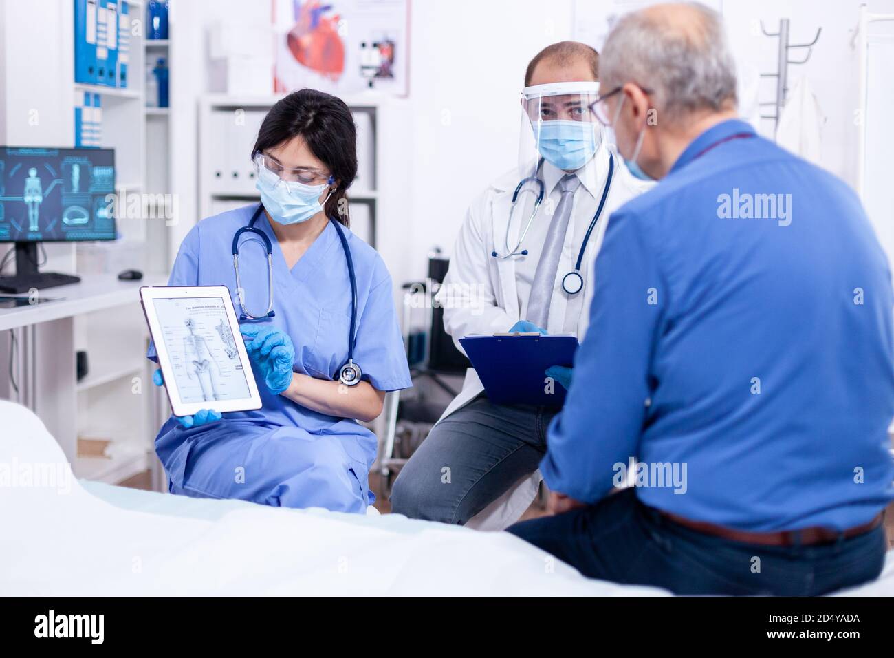 Medico e infermiere che spiegano la radiografia all'uomo anziano durante la consultazione sul pc della compressa in ospedale con protezione mas contro covid-19. Visita medica per infezioni, malattie e diagnosi. Foto Stock