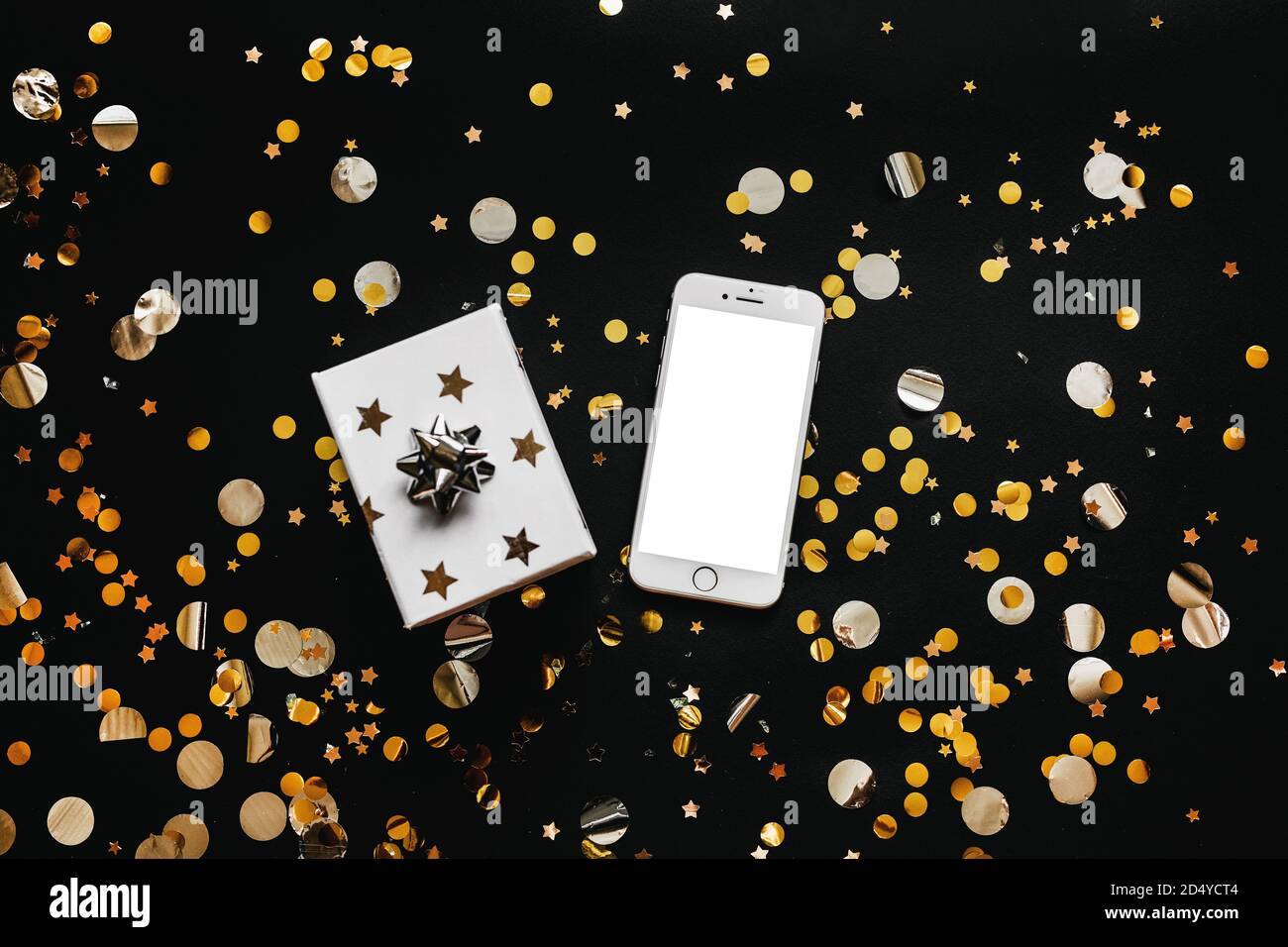 Telefono cellulare con schermo bianco vuoto o spazio di copia o mockup e accanto a una scatola con un regalo su sfondo nero con confetti d'oro. Background concettuale della vendita delle vacanze. Foto Stock