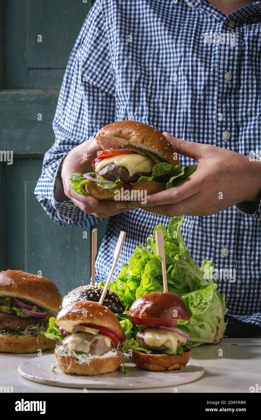 Uomo in camicia blu tenendo in mano in casa Hamburger classico nel frumento bun con costolette di manzo, formaggio fuso, verdure. Tavolo con ingredienti e mini Foto Stock