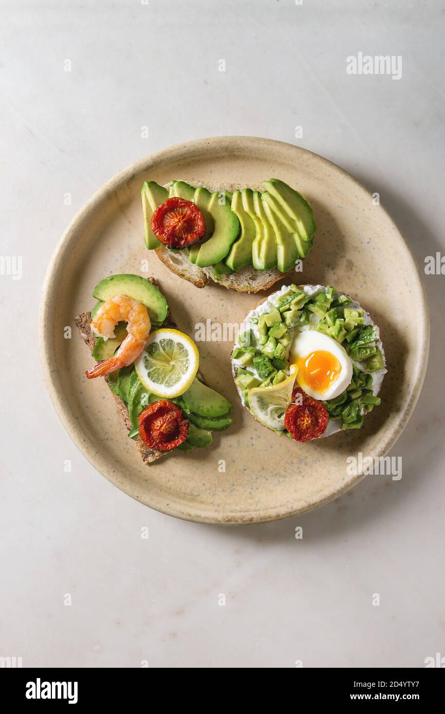 Varietà di panini vegetariani con fette di avocado, pomodori secchi, uovo, gamberetti, rucola servita su piastra in ceramica oltre bianco grigio maculato backgr Foto Stock