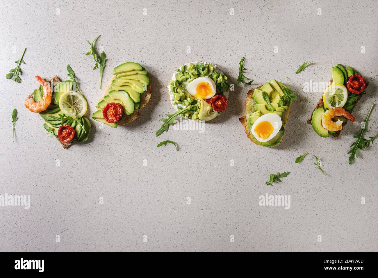 Varietà di panini vegetariani con fette di avocado, pomodori secchi, uovo, gamberetti, rucola servita su bianco grigio Sfondo maculato. Piatto, laici s Foto Stock