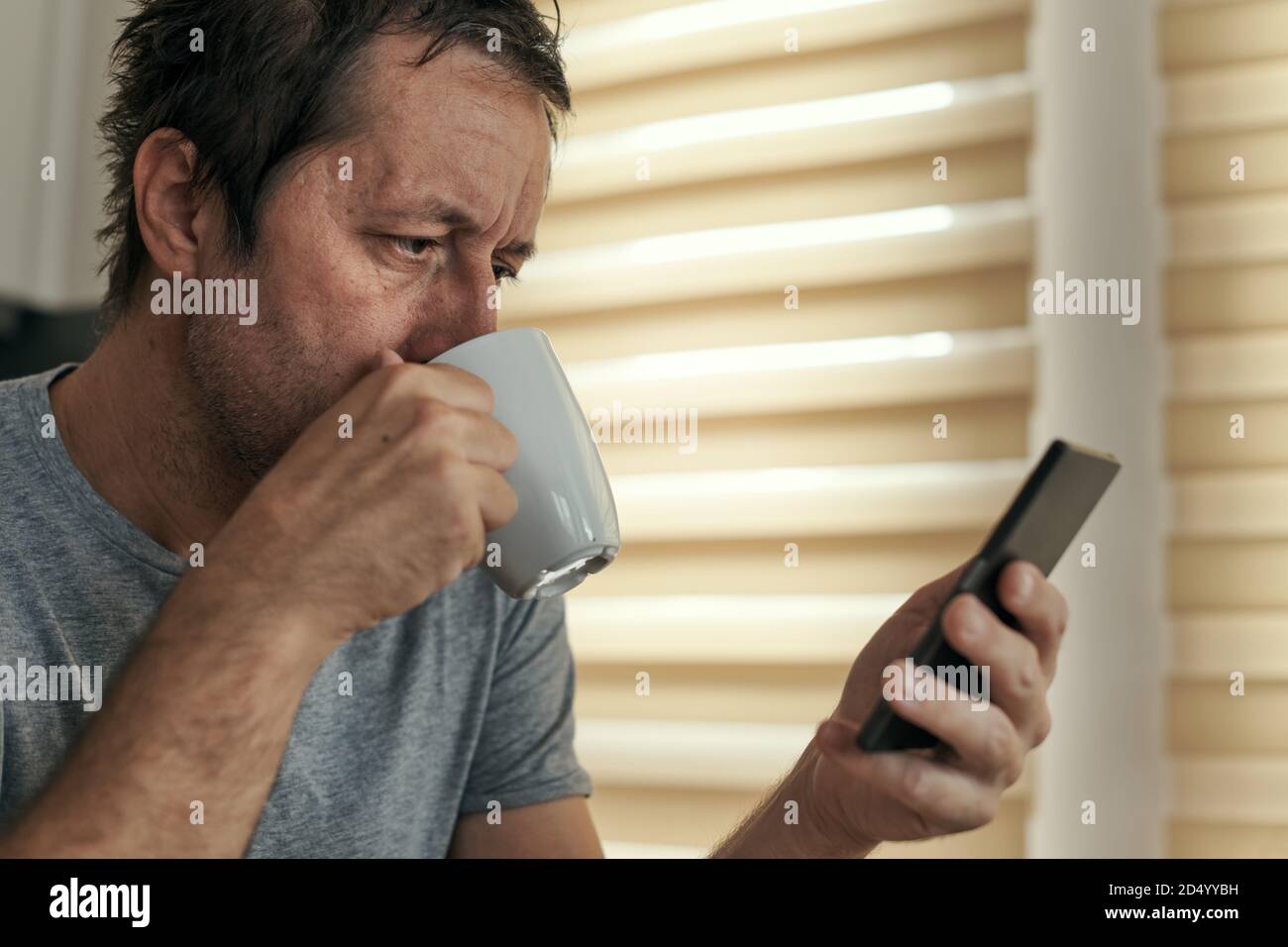 Uomo che beve caffè e utilizzando il telefono cellulare a casa, adulto maschio caucasico godendo di routine mattutina Foto Stock