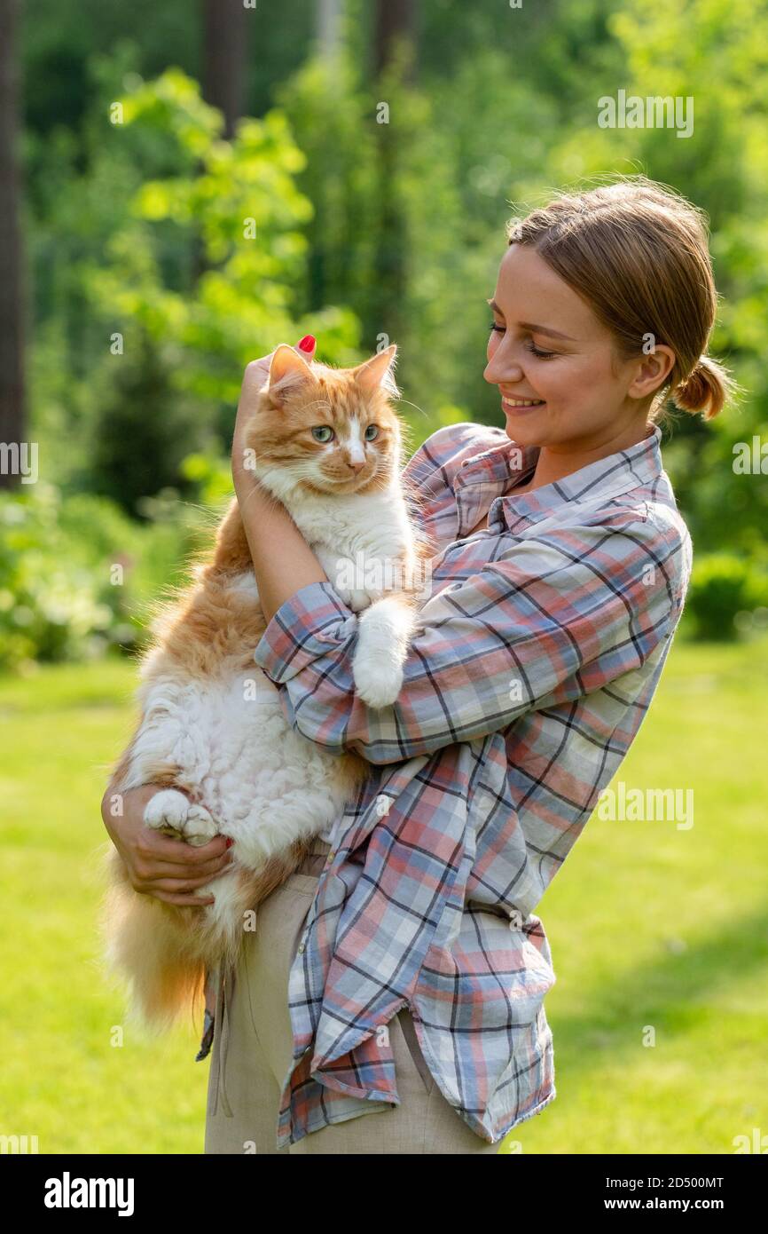 Primo piano di donna sorridente in camicia controllata guardando la macchina fotografica, abbracciando e abbracciando con tenerezza e amore domestico zenzero gatto all'aperto. Amore per Foto Stock