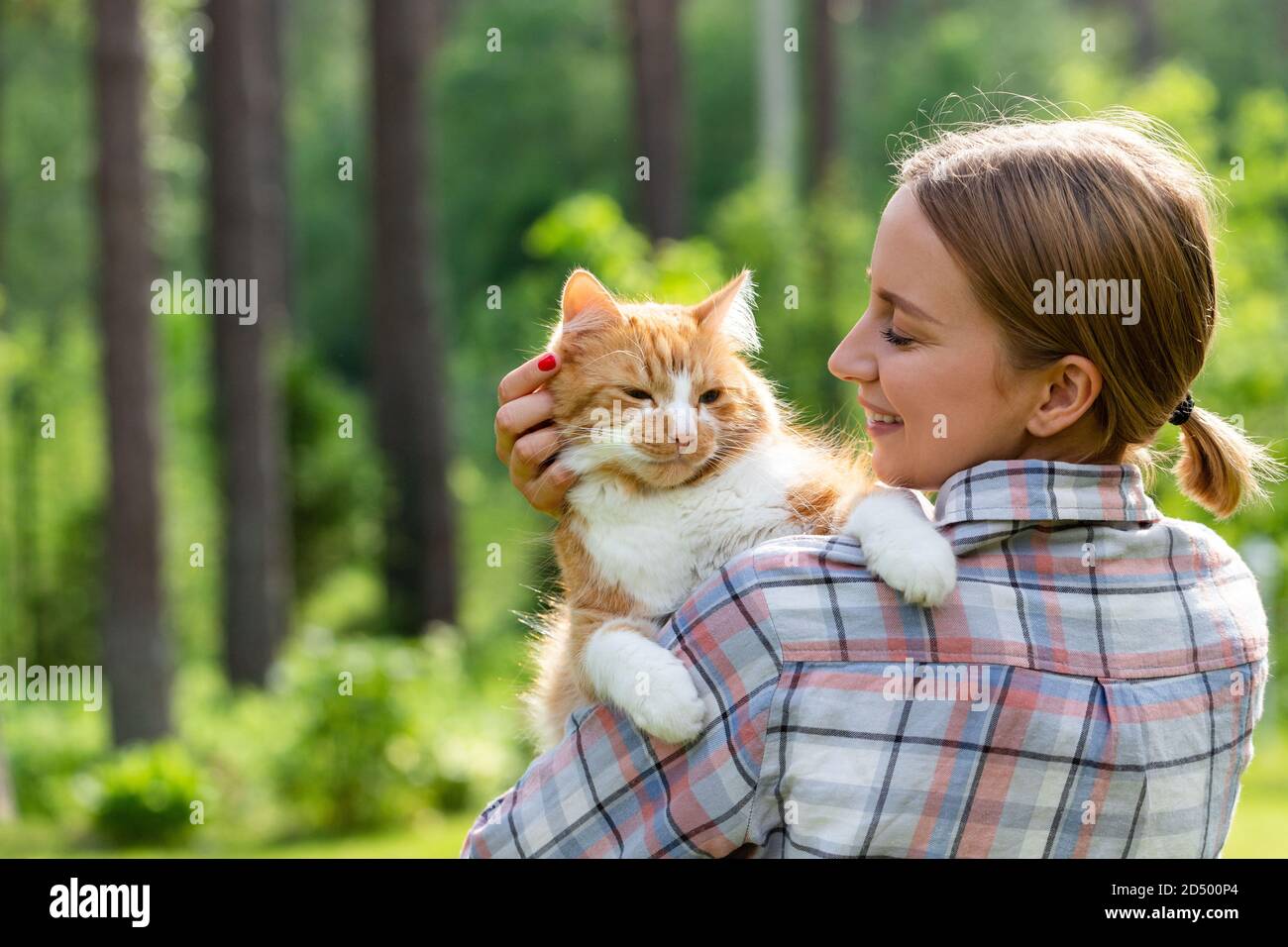 Primo piano di donna sorridente in camicia controllata abbracciando e abbracciando con tenerezza e amore gatto zenzero domestico, struggendo sulla testa, all'aperto in sole Foto Stock